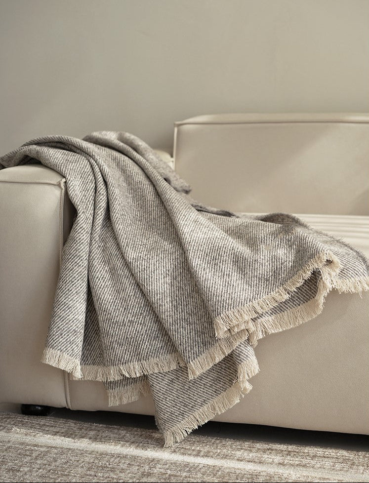 Grijze beige plaid met een subtiel patroon, gedrapeerd over de arm van een beige bank, met franjes aan de onderkant voor een elegante afwerking.