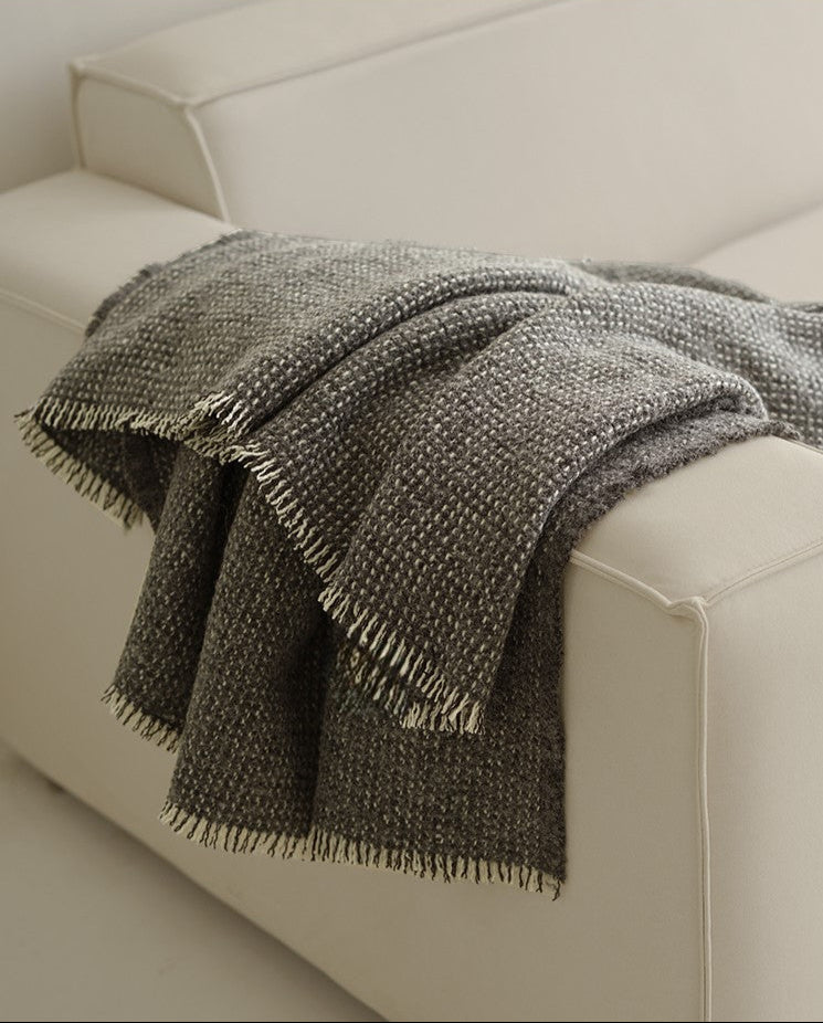 Grijze angora-wol en wollen plaid met franjes, elegant over de hoek van een lichte bank gedrapeerd in een moderne woonkamer.