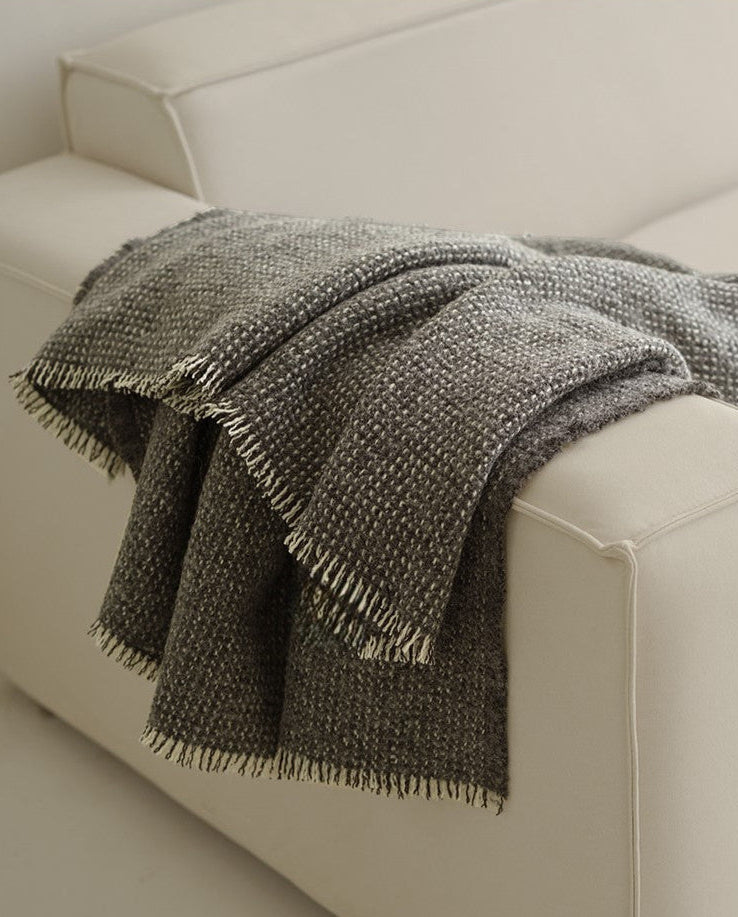 Grijze angora-wol en wollen plaid met franjes, elegant over de hoek van een lichte bank gedrapeerd in een moderne woonkamer.
