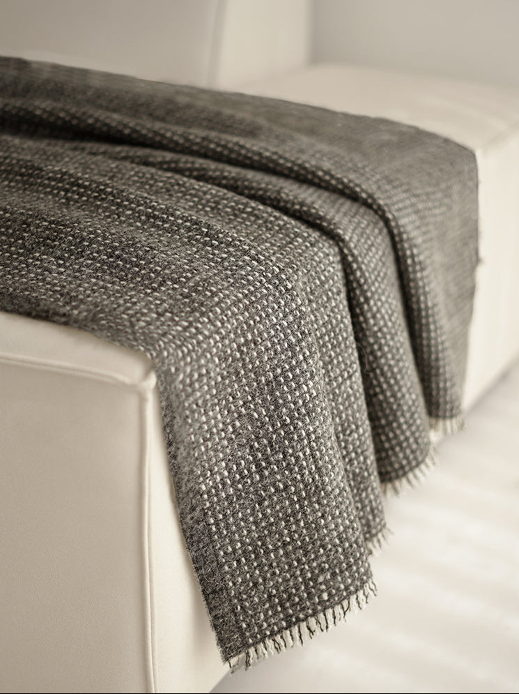 Grijze angora-wol en wollen plaid met franjes, subtiel over de armleuning van een moderne witte bank gedrapeerd.