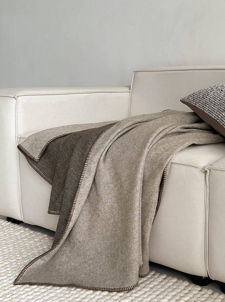 Two-tone deken in warme beige tinten, ontspannen over de bank gedrapeerd, met een luxueuze randafwerking, gecombineerd met een bijpassend kussen.