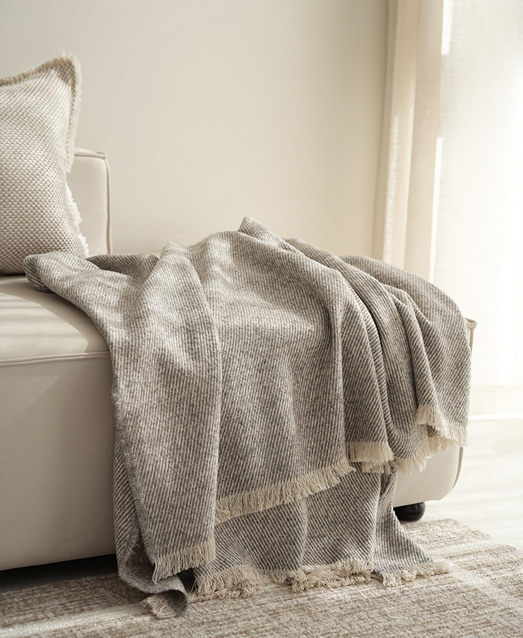Grijze beige plaid met een subtiel patroon, gedrapeerd over de arm van een beige bank, met franjes aan de onderkant voor een elegante afwerking.