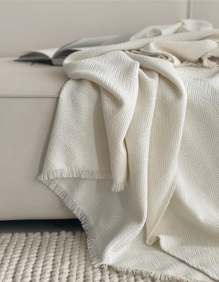 Classic witte Herringbone plaid met franjes op de bank, gemaakt van luxe wolmix, met een subtiel visgraatpatroon en een boek op de achtergrond.