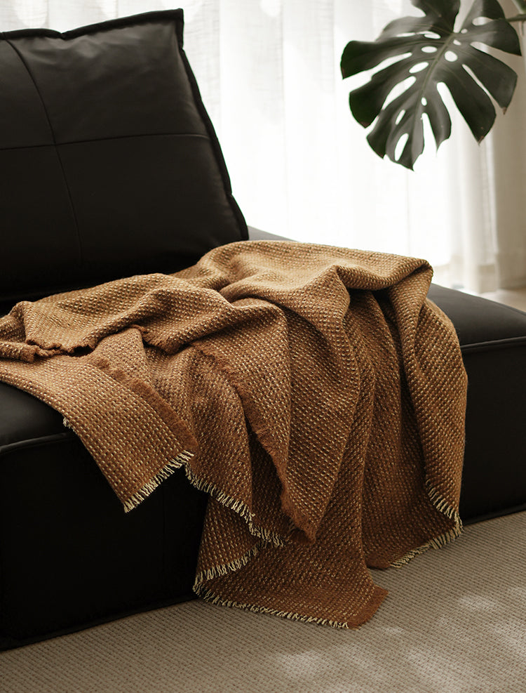 Luxe angora wol en wollen plaid in bruine kleur, met franjes, draperend over een zwarte bank in een stijlvolle woonkamer met planten.