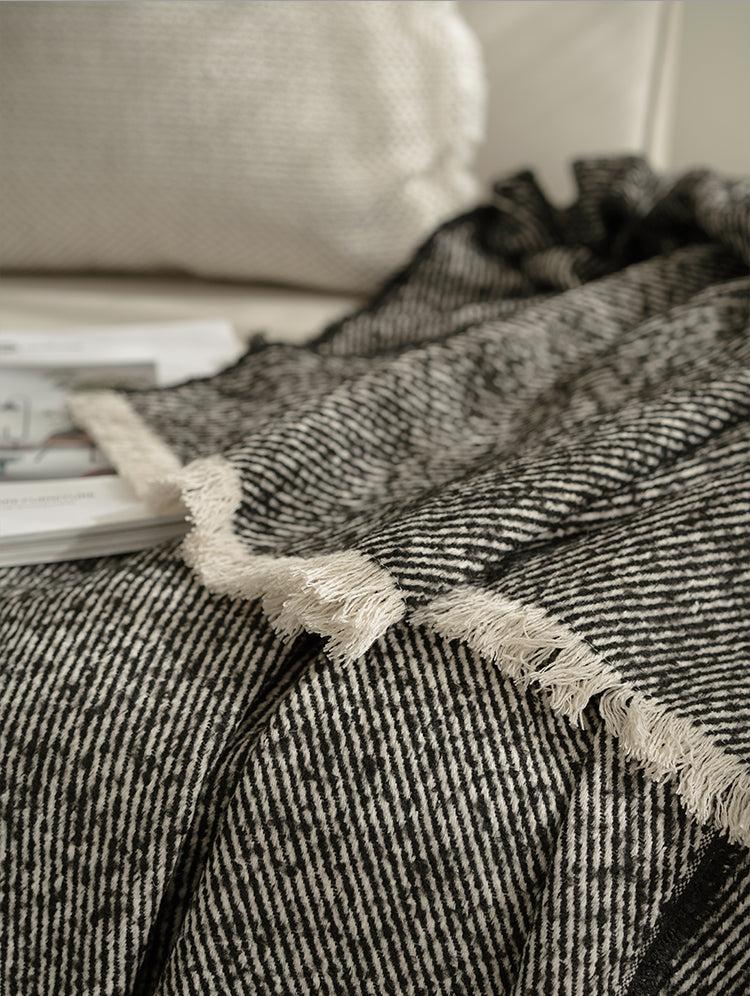 Close-up van een zwarte plaid met een gestreept patroon, liggend op een bank, met een boek ernaast en kussen op achtergrond