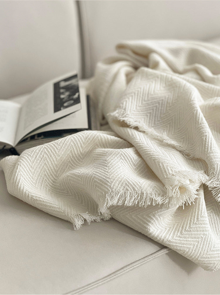 Classic witte Herringbone plaid met franjes op een lichte bank, met een open boek ernaast, gemaakt van luxe wolmix met een visgraatpatroon.