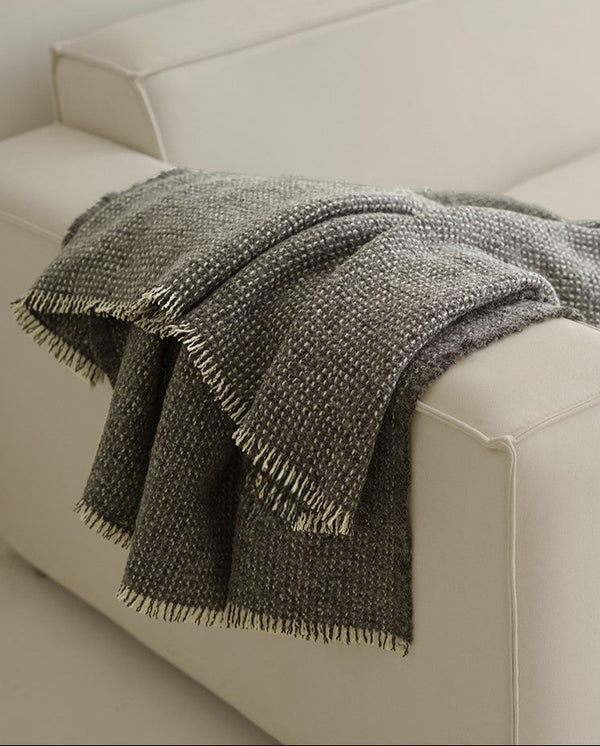 Grijze angora-wol en wollen plaid met franjes, elegant over de hoek van een lichte bank gedrapeerd in een moderne woonkamer.